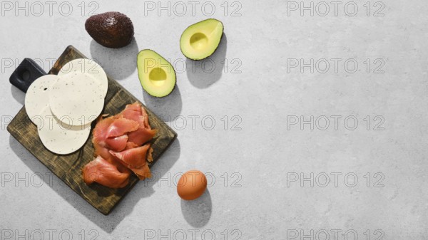Smoked salmon, slices of goat cheese, halved avocados, and an egg on a wooden cutting board. Healthy food ingredients