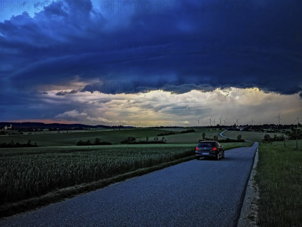 Supercell, car driving on a small road towards an approaching thunderstorm, impressive and threatening cloud formation of a thunderstorm cell above a village with wind turbines in the background, Baden-Württemberg, Germany