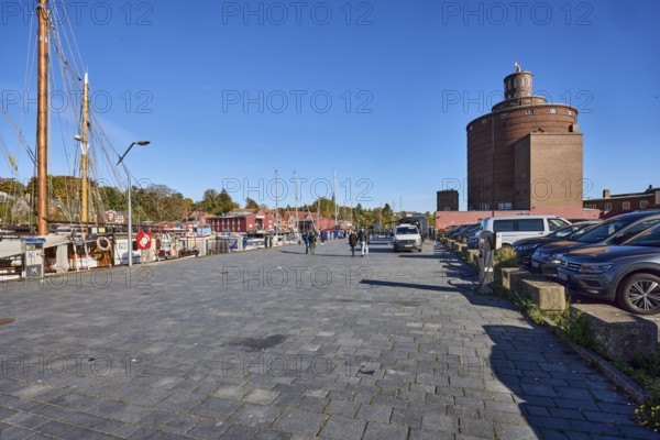 Round silo, historic brick building, architect Heinrich Hansen, architectural style New Objectivity, harbor, wharf, ships, street, lantern, parking boxes with cars, trees, blue sky, cirrus clouds, Hafenstraße, Eckernförde, Rendsburg-Eckernförde district, Schleswig-Holstein, Germany