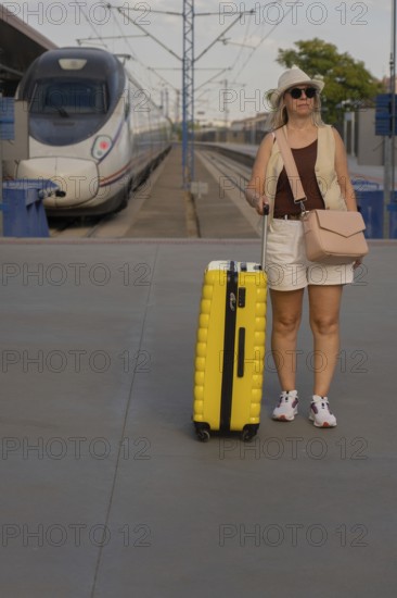 Adult female traveler standing on a railway platform with a yellow suitcase, looking around while waiting for her train in a modern city station