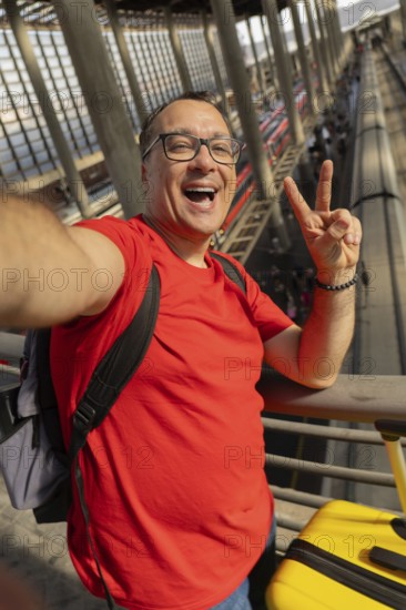 Middle aged man wearing reading glasses smiles with teeth while taking a selfie and making a peace sign at a modern train station. Positive travel mood and urban lifestyle