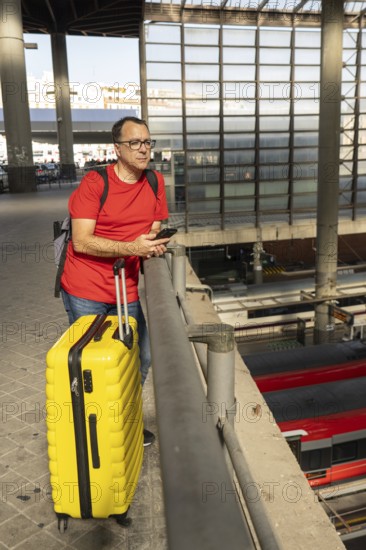 Middle aged man standing at a city train station with a yellow suitcase and looking at the railway tracks below during an urban travel scene