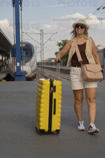 Adult woman walking alone on a city train platform, pulling a yellow suitcase and looking ahead while waiting for public transportation in a modern urban environment