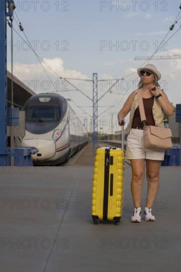 Adult woman walking on a modern train platform with a yellow suitcase, waiting for departure. Solo travel, urban transport, summer vacation lifestyle in Europe
