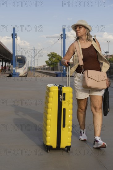 Adult woman traveling alone at a city train station, pulling a yellow suitcase while walking along the platform during a relaxed daytime journey