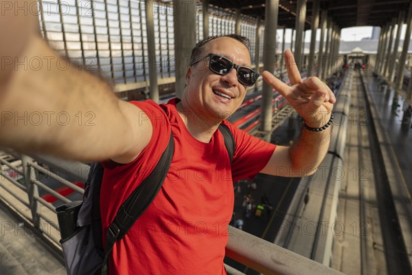 Middle aged man wearing sunglasses takes a cheerful selfie at a busy train station. Modern travel, urban lifestyle, summer journey and vacation concept in Europe