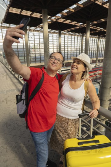 Smiling adult travel couple taking a selfie and looking at a smartphone while standing inside a modern train station with luggage. Selfie travel moment, tourism, transportation and lifestyle concept