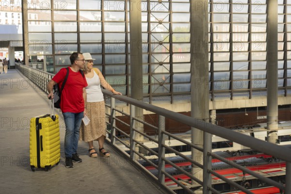 Smiling adult travel couple standing with luggage on an upper level of a modern train station, looking down at the trains below. The woman wears a hat, the man wears a red t shirt and holds boarding passes. Multimodal transport, travel connection, tourism and lifestyle concept