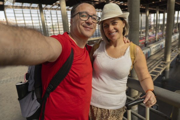 Happy adult travel couple taking a selfie and looking at the camera while standing inside a modern train station with luggage. Travel photography, tourism, transportation and lifestyle concept