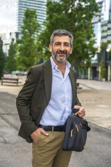 Confident mature businessman with beard and suit jacket smiling at camera, holding a messenger bag while standing in a modern urban street, projecting success and approachability