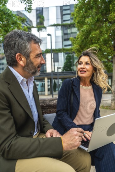 Two professional business colleagues, a man and a woman, are sitting on a bench in a city environment, collaborating and having an informal discussion while using a laptop