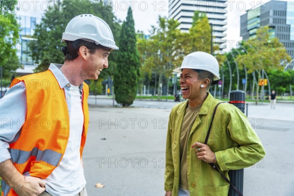 Two diverse construction professionals wearing hard hats and safety gear, happily collaborating and discussing plans on a street in an urban environment during the day