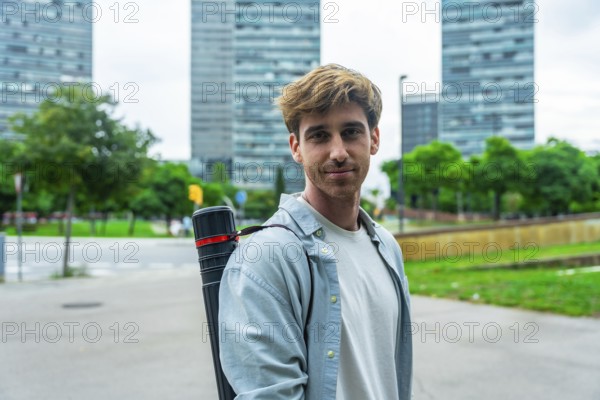 Young male architect standing outdoors with a blueprint tube, smiling confidently at the camera amid urban buildings and green trees, projecting ambition and modern city life