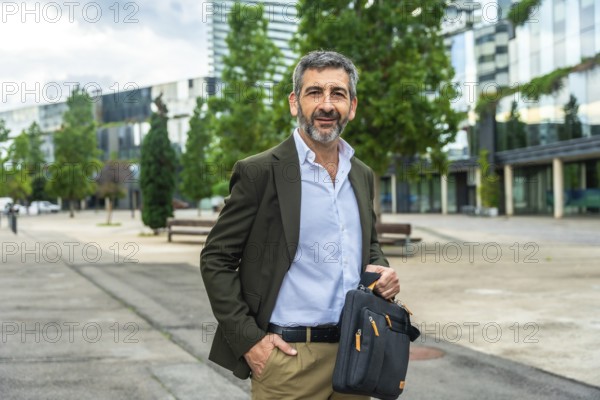 Confident mature businessman carrying a briefcase and smiling at the camera, standing outdoors with one hand in his pocket against a modern urban background, representing success and professionalism