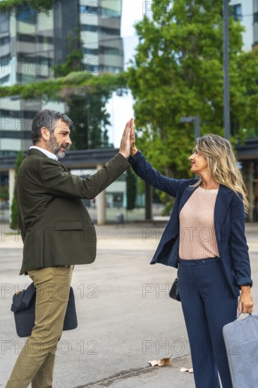 Two mature business professionals celebrating their teamwork and success with a high five, standing outdoors in an urban environment with office buildings in the background