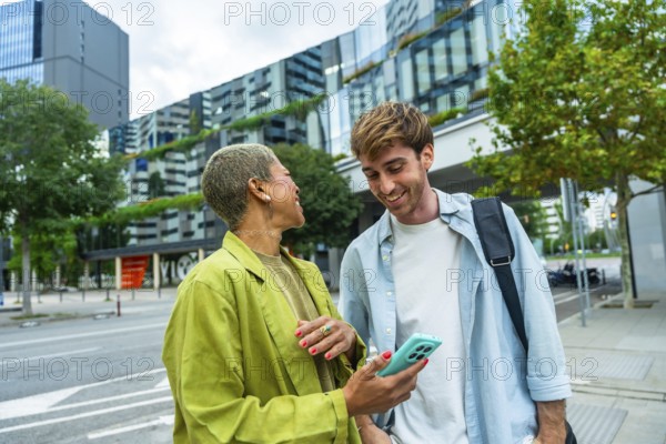 Two diverse colleagues enjoying a casual conversation and sharing a smartphone screen on an urban street, featuring modern city architecture and green spaces