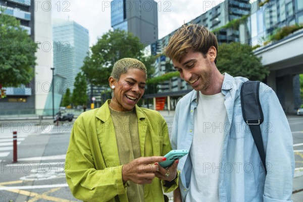 Diverse colleagues standing outdoors, laughing happily while looking at a smartphone, sharing a lighthearted moment together in a modern urban environment with city buildings