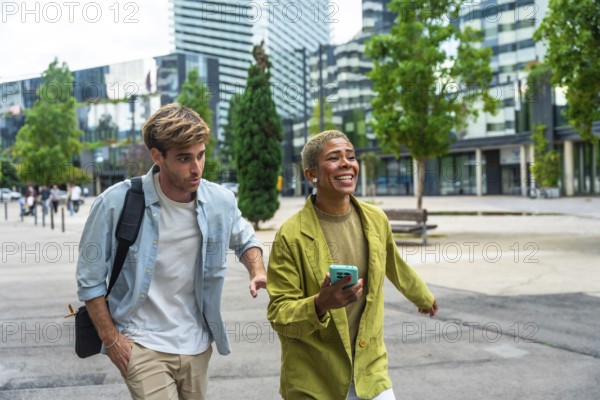 Diverse business colleagues walking on a paved pathway outdoors in a modern city district, sharing a joyful moment and communicating while carrying a shoulder bag and smartphone