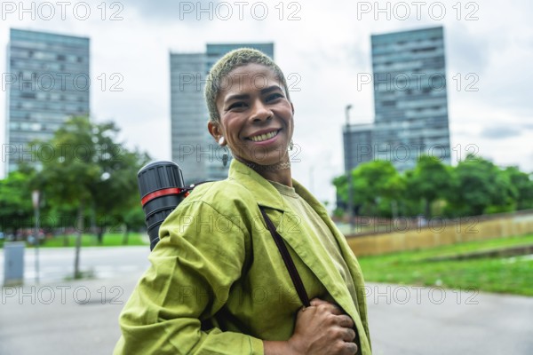 Urban architect smiling confidently with drawing tube on shoulder strap, standing in front of modern downtown skyscrapers, conveying creative success, ambition and forward thinking design