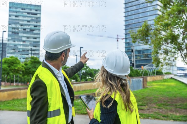 Architects and engineers wearing hard hats and safety vests discussing city development while pointing at a new construction project and modern skyscrapers