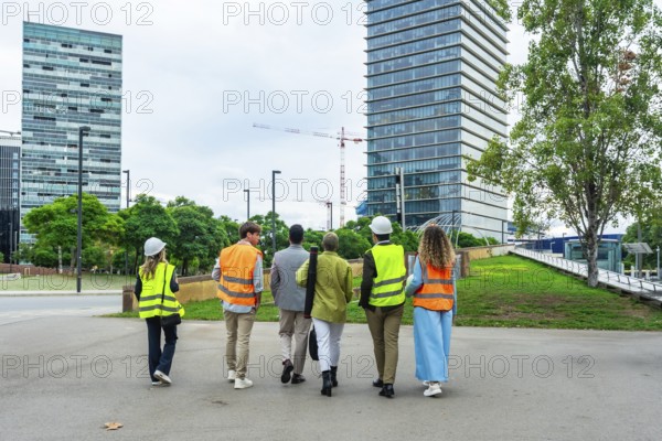 Diverse team of construction professionals and architects wearing safety vests and hard hats walking through a corporate park near modern buildings and a construction site