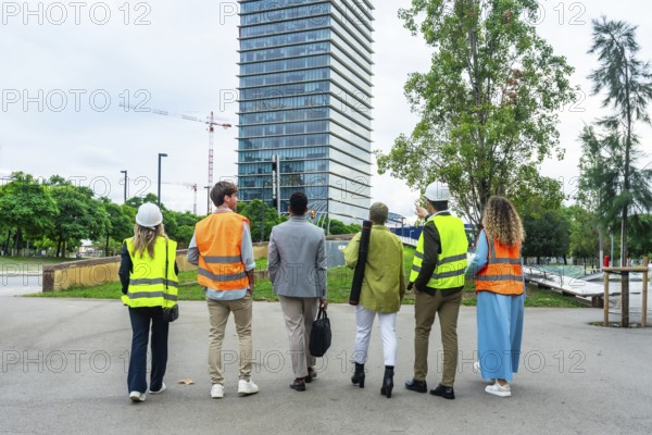 Diverse team of engineers, architects, and business executives standing with backs to camera, discussing the progress of a large construction project in a dynamic urban environment