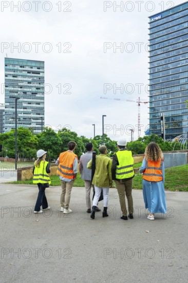 Group of executives and architects wearing safety vests and hard hats, observing a construction site with modern skyscrapers, discussing progress and future urban development plans