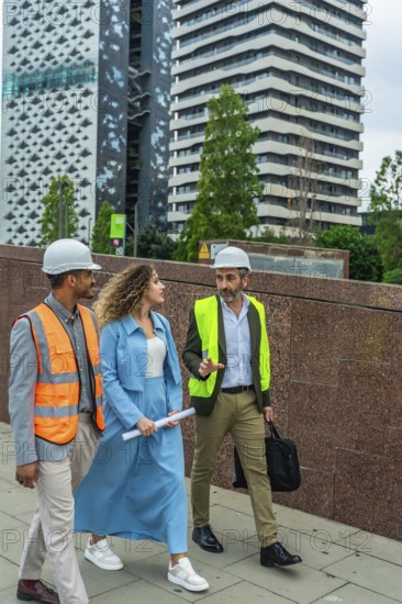 Construction professionals walking and discussing a project on a city street, wearing hard hats and safety vests while carrying blueprints and a briefcase