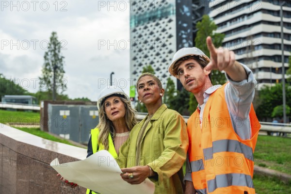Diverse construction team in hard hats and safety vests reviews blueprints and points toward an urban development site, collaborating on planning and inspection of future high rises