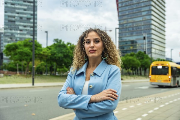 Young professional businesswoman with curly hair standing confidently with crossed arms on an urban street, featuring modern buildings, green trees, and a passing bus in the background