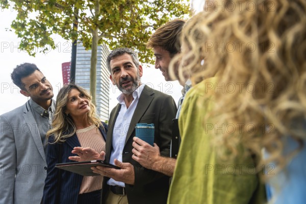 Diverse group of business professionals meeting outdoors, discussing data on a digital tablet, fostering teamwork and urban collaboration among entrepreneurs