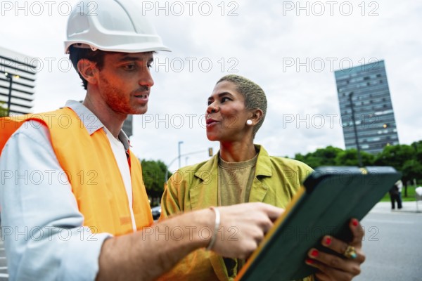 Construction engineer wearing a hard hat and safety vest pointing to a digital tablet while having a conversation with a colleague in an urban environment