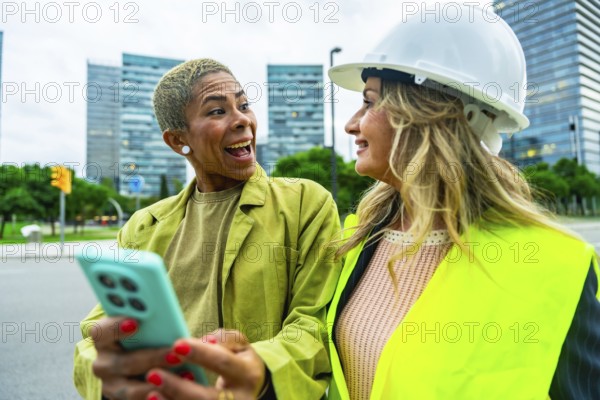 Two diverse colleagues. One an architect wearing a hardhat and safety vest. Looking at a smartphone held by her associate. Sharing a laugh while working together on a construction project in the city