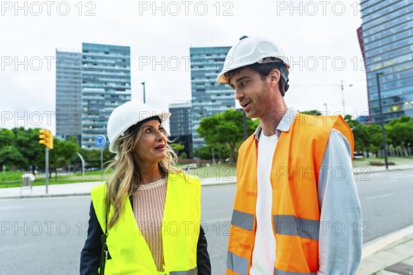 Two professionals, one woman and one man, wearing hard hats and safety vests, standing on a city street and conversing about a project with modern buildings in the background