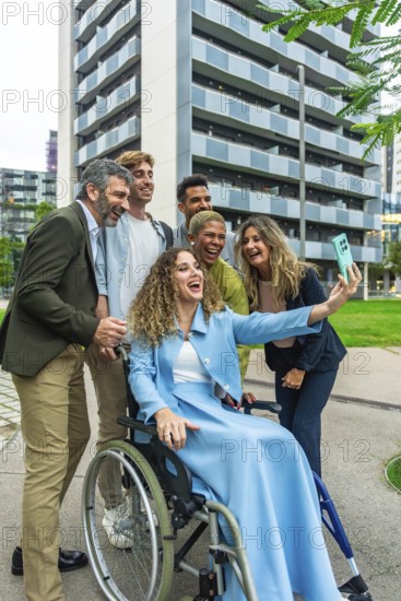 Diverse group of smiling colleagues with a woman in a wheelchair taking a cheerful selfie together outdoors in an urban area, demonstrating teamwork and inclusion