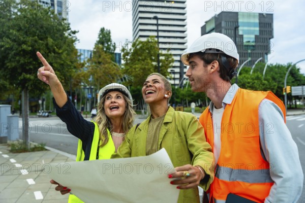 Diverse team of architects and engineers smiling and pointing towards a building, holding blueprints and discussing urban development plans outdoors in the city