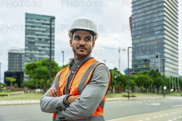 Young man wearing a hard hat and safety vest standing confidently with arms crossed in a cityscape, representing construction, urban development, and professional expertise