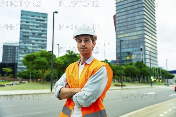 Confident man wearing a hard hat and safety vest standing with crossed arms in a modern urban area, representing construction, engineering, and architecture professions