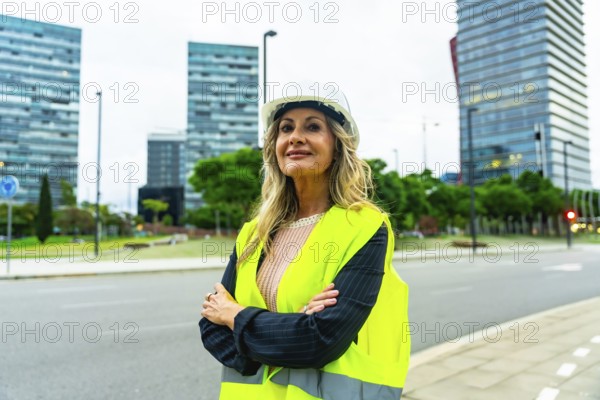 Confident mature professional woman in a hard hat and safety vest standing with crossed arms in a modern urban environment, representing leadership and expertise in civil engineering or construction