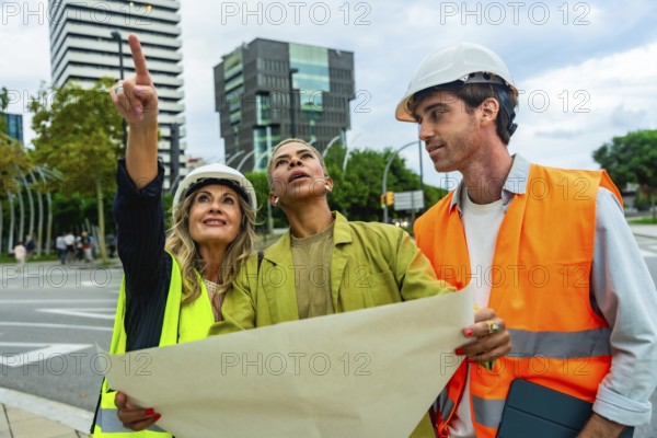 Diverse team of architects and engineers wearing hard hats and safety vests, examining blueprints and discussing urban development while pointing at a building site in a modern city