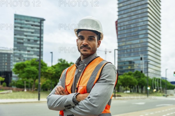 Professional engineer or architect standing confidently with crossed arms, wearing a hardhat and high visibility vest with modern city buildings and urban landscape in the background