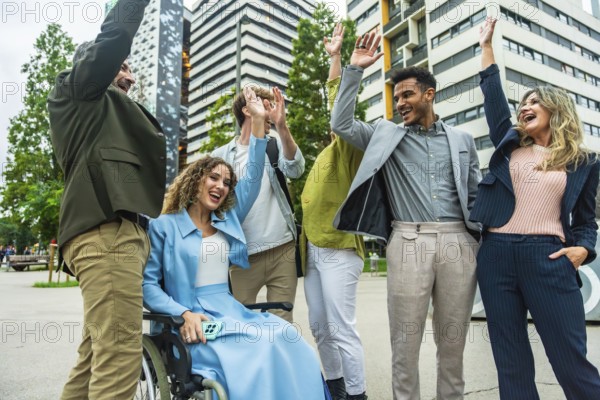 Diverse business colleagues including a woman in a wheelchair cheering and doing high fives outdoors in a modern city, celebrating teamwork, inclusion, accessibility, and achievement