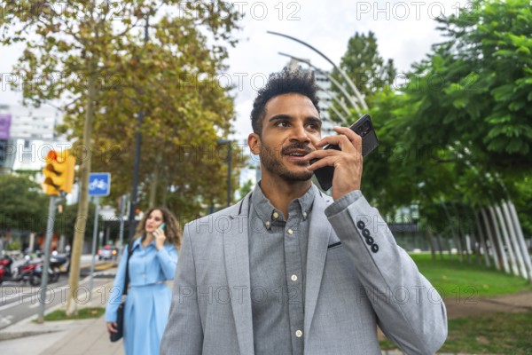 Confident young business professional communicating on a mobile phone, using its speaker function as he walks along a green urban sidewalk in a modern city