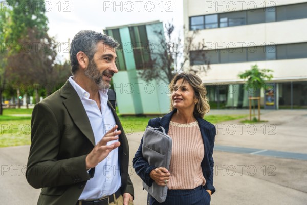 Two business colleagues, a man and a woman, walking together in an urban park setting while engaging in a professional conversation, both dressed in smart casual attire