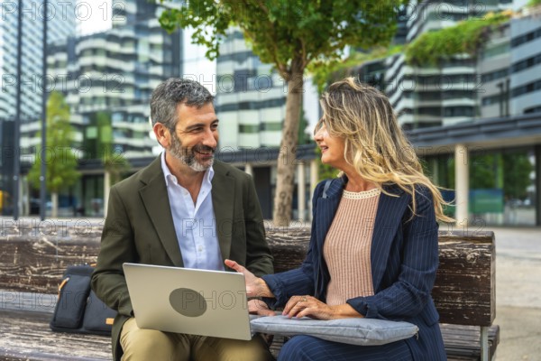 Two smiling business colleagues having an informal outdoor meeting on a bench, collaborating on a project using a laptop in a modern city setting with corporate buildings in the background