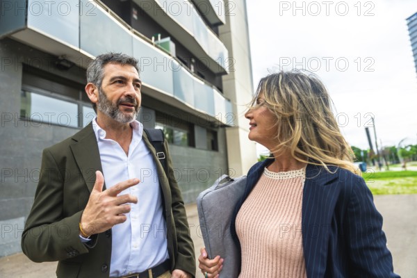 Two business colleagues, a man and a woman, are walking and having a professional discussion outside a modern building in an urban environment, engaging in communication and collaboration