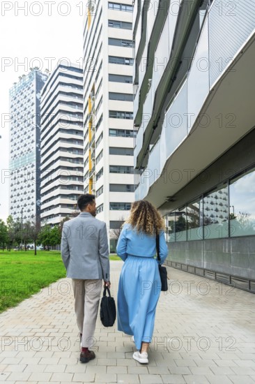Two business professionals walking together along a paved pathway in an urban area, passing tall contemporary buildings, representing teamwork, partnership, and city life