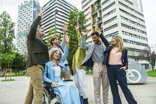 Mixed age businesspeople with a woman in a wheelchair raising their hands in celebration, showing unity, teamwork, and inclusion in a modern city setting with buildings and green space