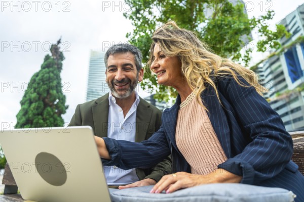 Business colleagues smiling and discussing work outdoors in a park, reviewing information on a laptop with modern city buildings and trees in the background