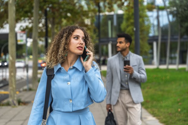 Confident businesswoman talking on her smartphone while walking on a city sidewalk with a male colleague carrying a laptop bag in the background, representing modern urban professionals